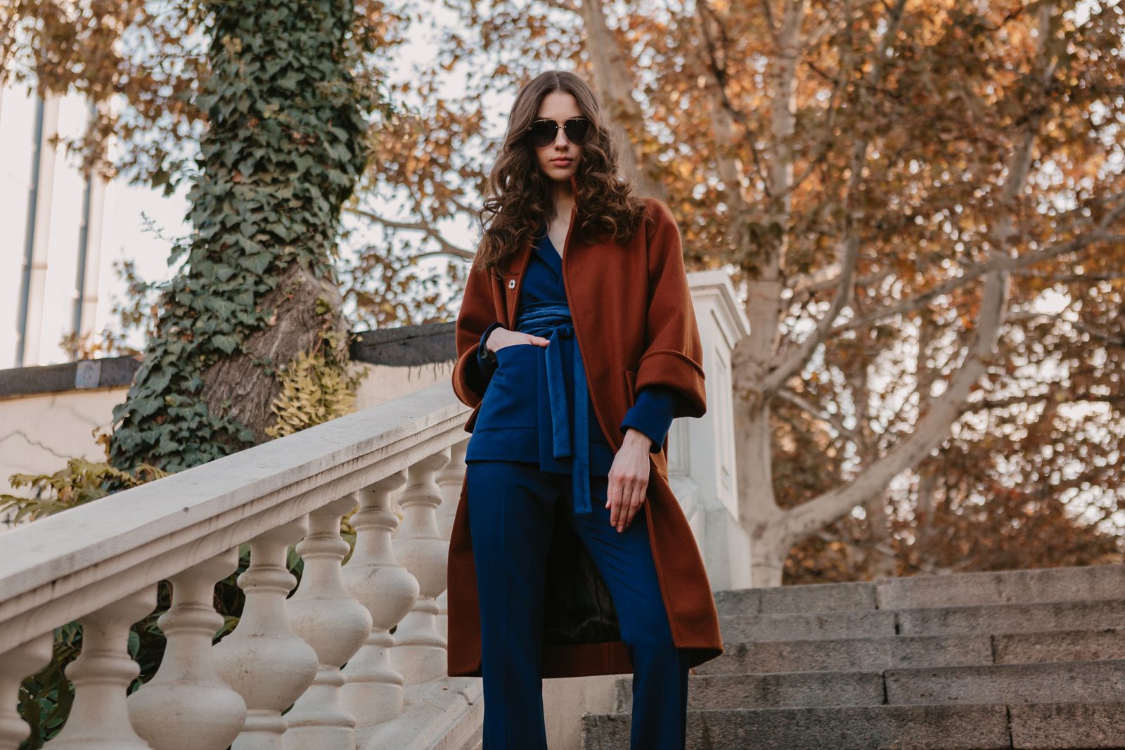 Žena v cihlovém kabátu a modrém outfitu na schodech